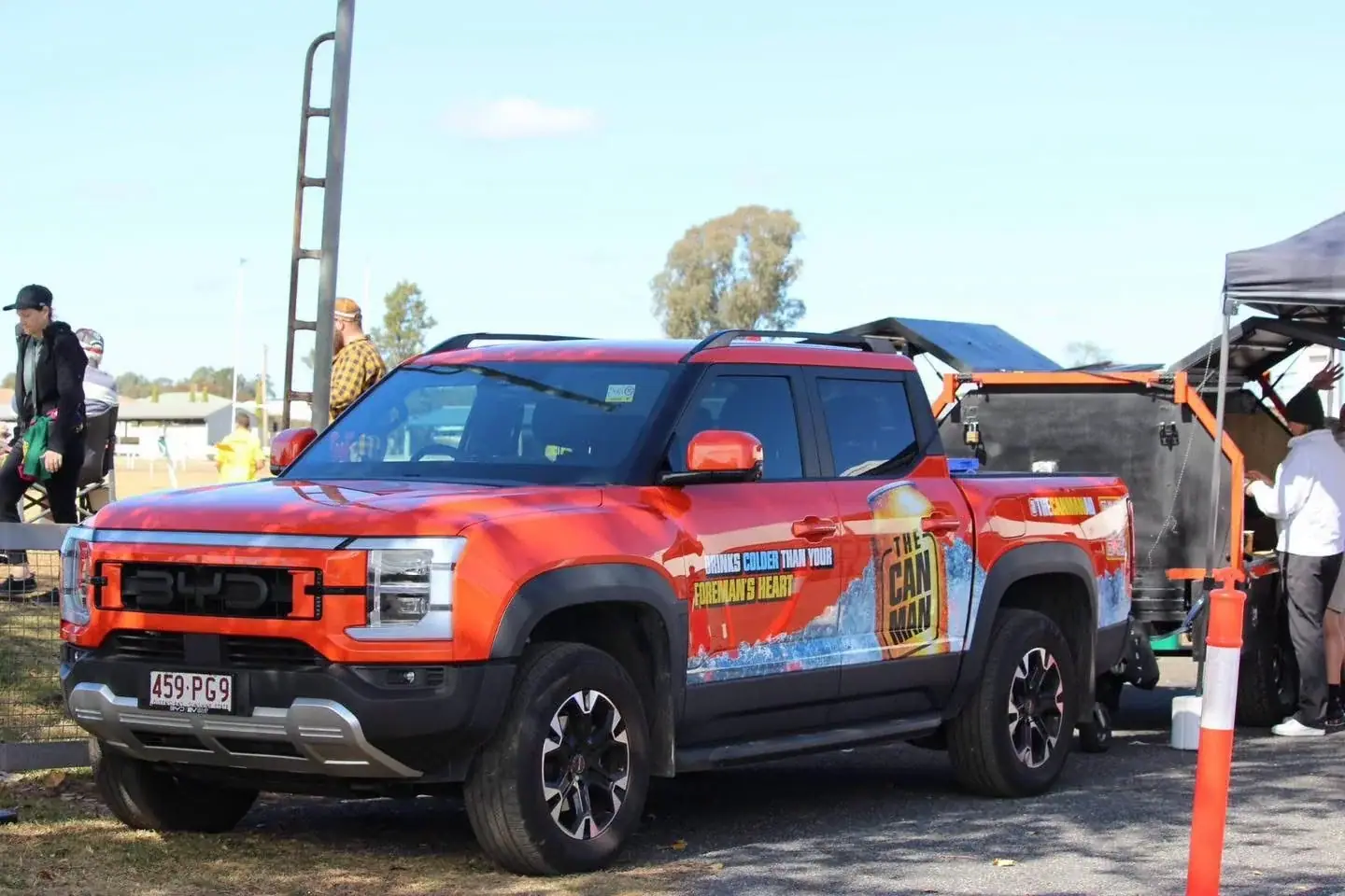 Red BYD Shark 6 ute with The Can Man livery towing the matte-black coffee trailer at a daytime sports and club event