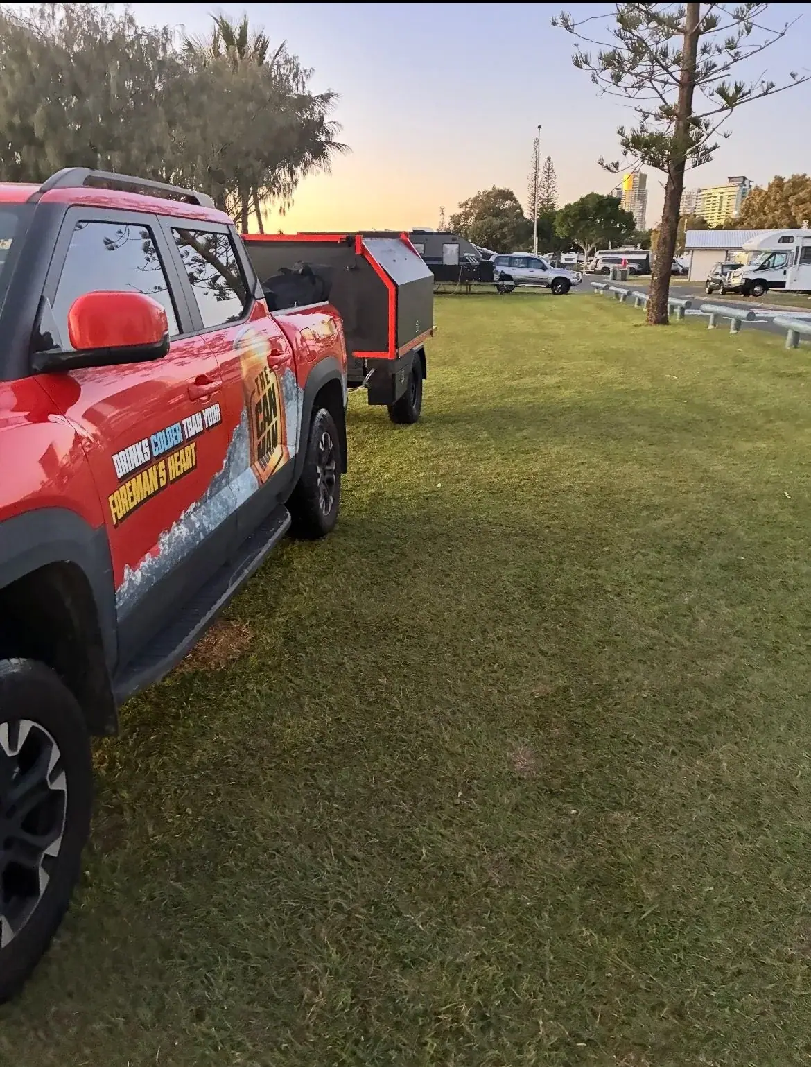 Red Can Man Coffee ute towing its matte-black trailer parked on grass beside the water at first light